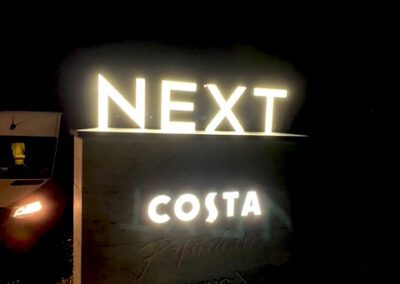 Freestanding face illuminated Next letters on top of stone clad totem with Costa letters